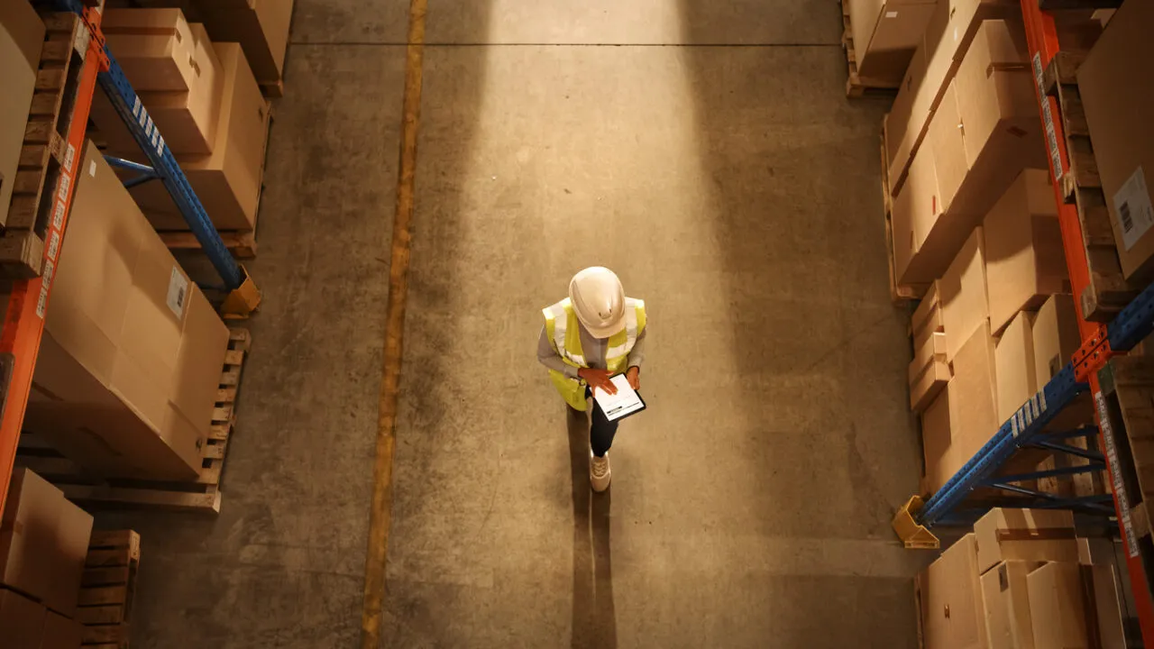 Top-Down View: Worker Wearing Hard Hat Checks Stock and Inventory Using Digital Tablet Computer in the Retail Warehouse full of Shelves with Goods. Working in Logistics, Distribution Top-Down View: Worker Wearing Hard Hat Checks Stock and Inventory Using Digital Tablet Computer in the Retail Warehouse full of Shelves with Goods. Working in Logistics, Distribution