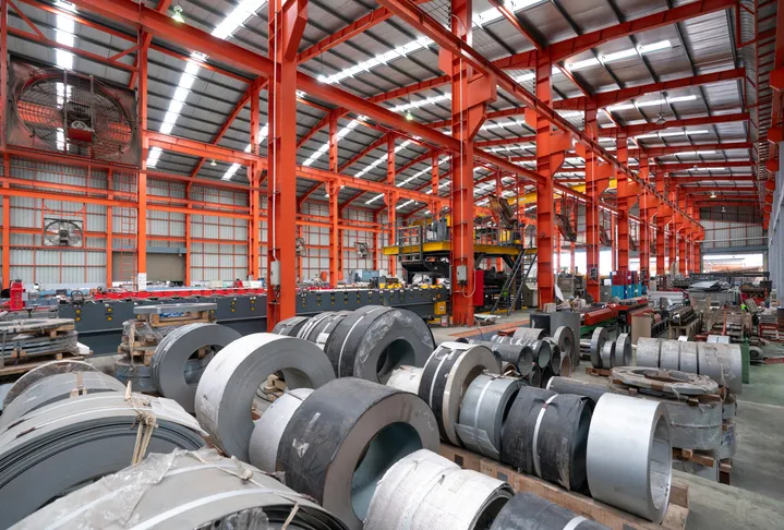 A gargantuan warehouse brims with a labyrinth of gleaming steel beams crisscrossing overhead, creating a metallic canopy. There are steel sheet roll and metal sheets on the floor. A gargantuan warehouse brims with a labyrinth of gleaming steel beams crisscrossing overhead, creating a metallic canopy. There are steel sheet roll and metal sheets on the floor.